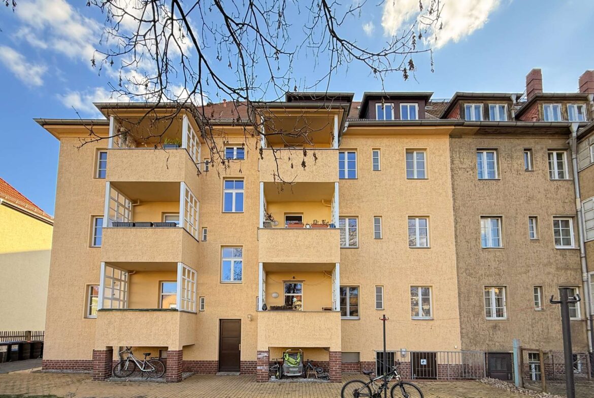 Beige, four-story apartment building with open balconies and bicycles parked at ground level against a blue sky with bare tree branches above.