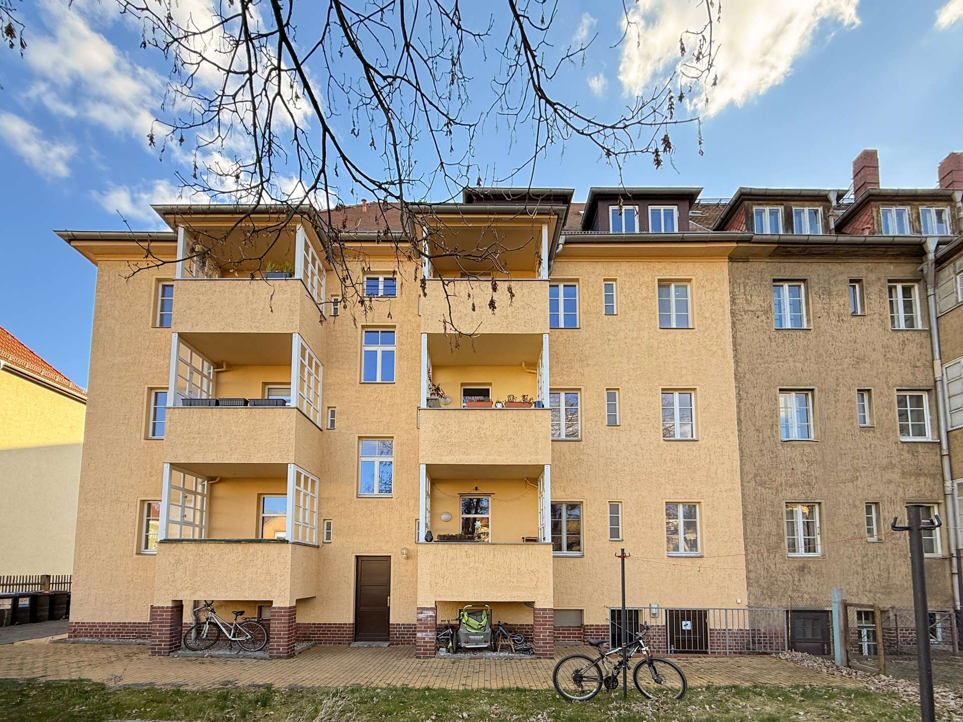 Beige, four-story apartment building with open balconies and bicycles parked at ground level against a blue sky with bare tree branches above.