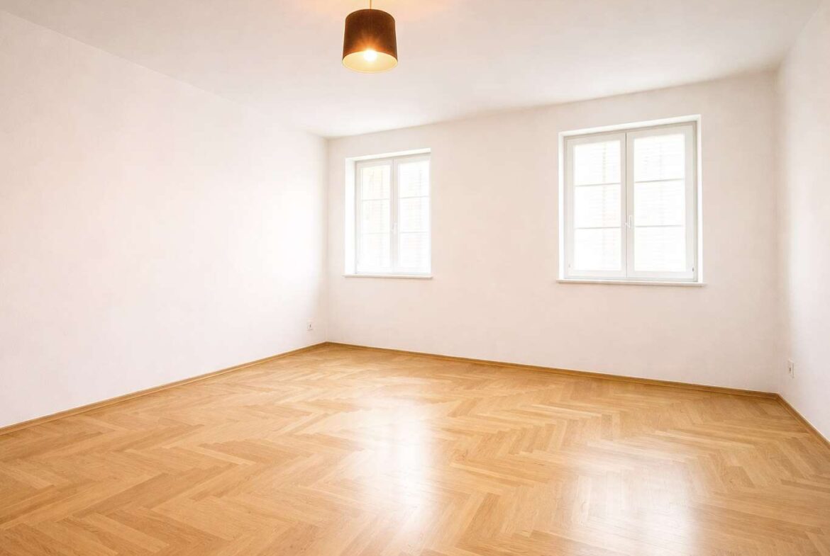 Empty bright room with light wood herringbone floor, white walls, two windows, and a single ceiling light fixture (unfurnished living space).