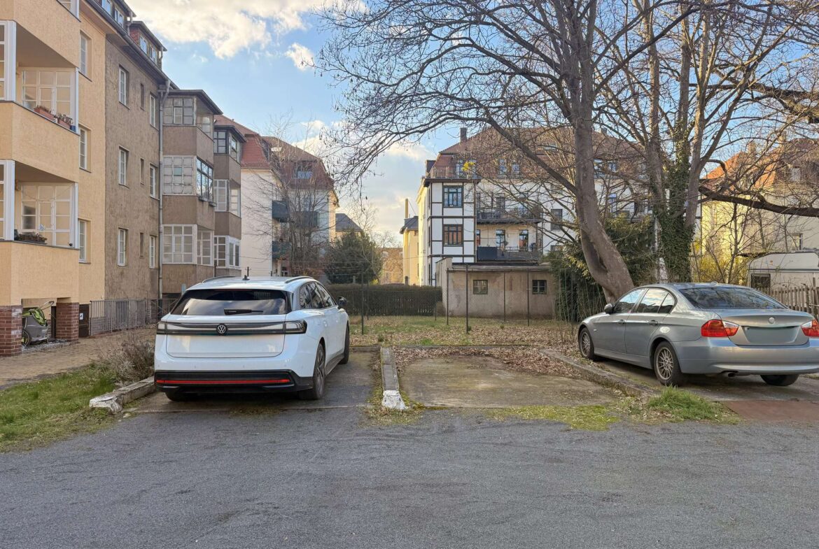 White SUV (VW) parked in a residential parking lot beside beige apartment buildings, with a large leafless tree in the background and blue sky above.