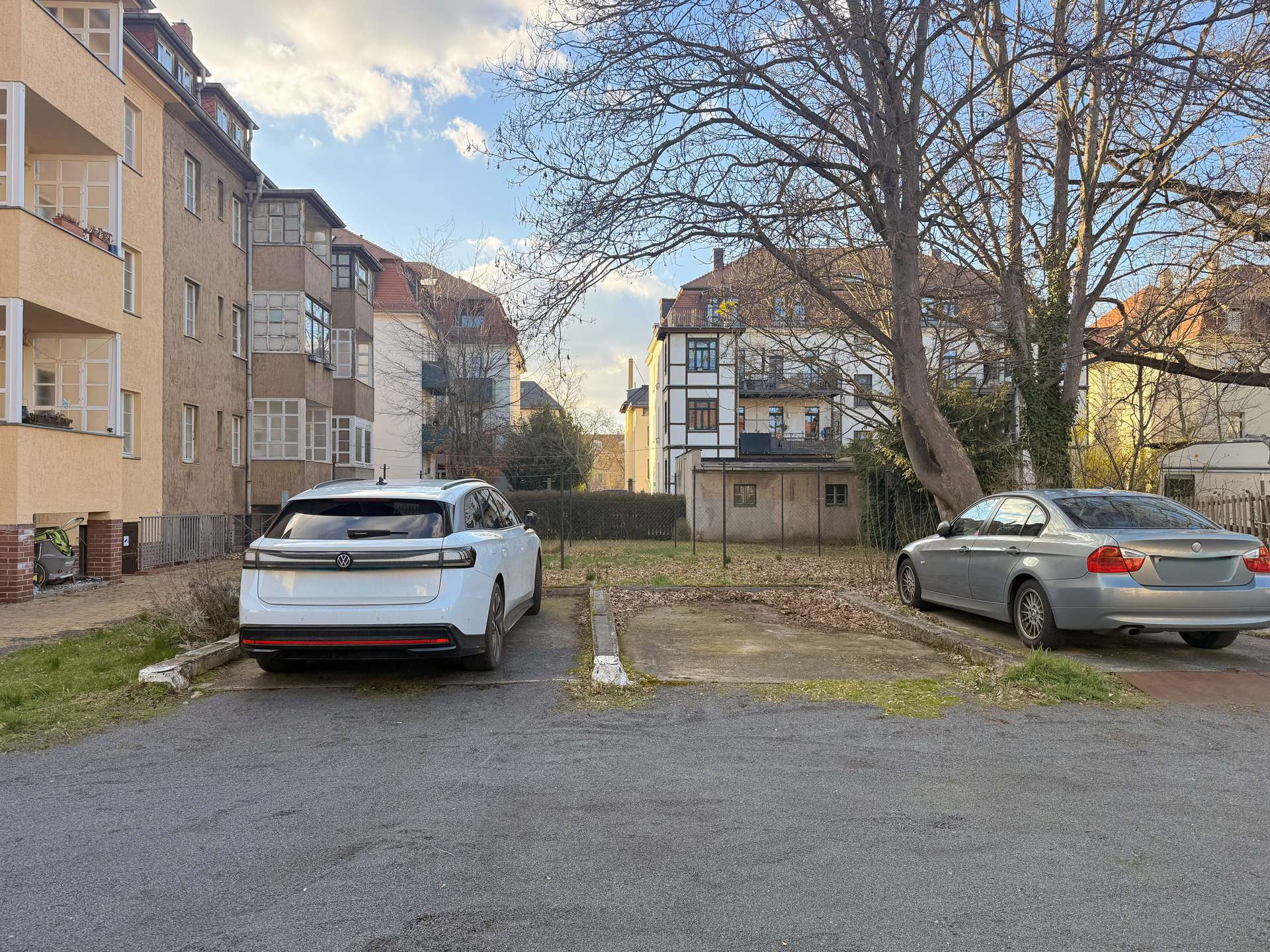 White SUV (VW) parked in a residential parking lot beside beige apartment buildings, with a large leafless tree in the background and blue sky above.