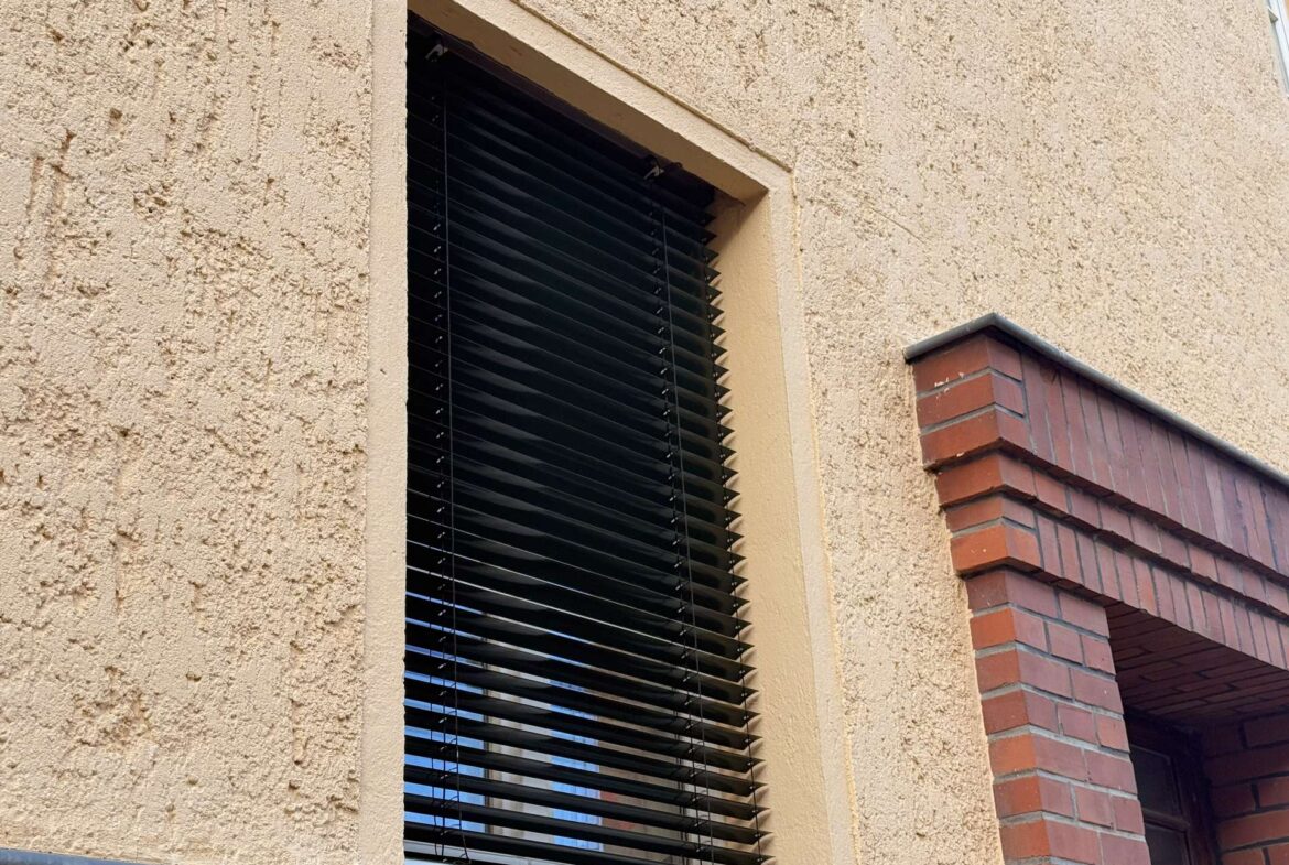 Exterior window with black horizontal blinds on a textured beige stucco wall, brick architectural feature to the right