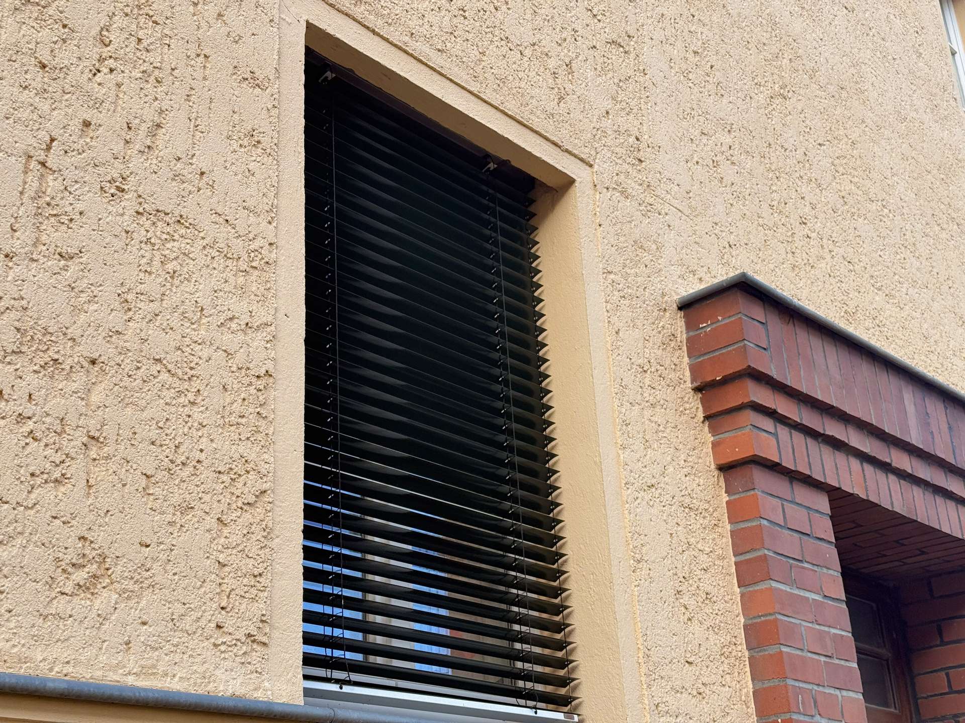 Exterior window with black horizontal blinds on a textured beige stucco wall, brick architectural feature to the right