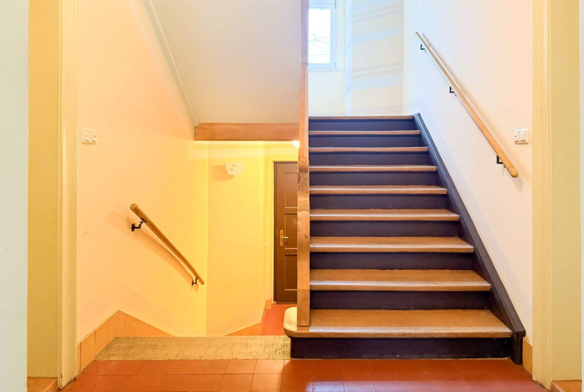 Staircase in a building: wooden steps with dark risers, beige walls, and handrails on both sides, leading to a door and a window above.
