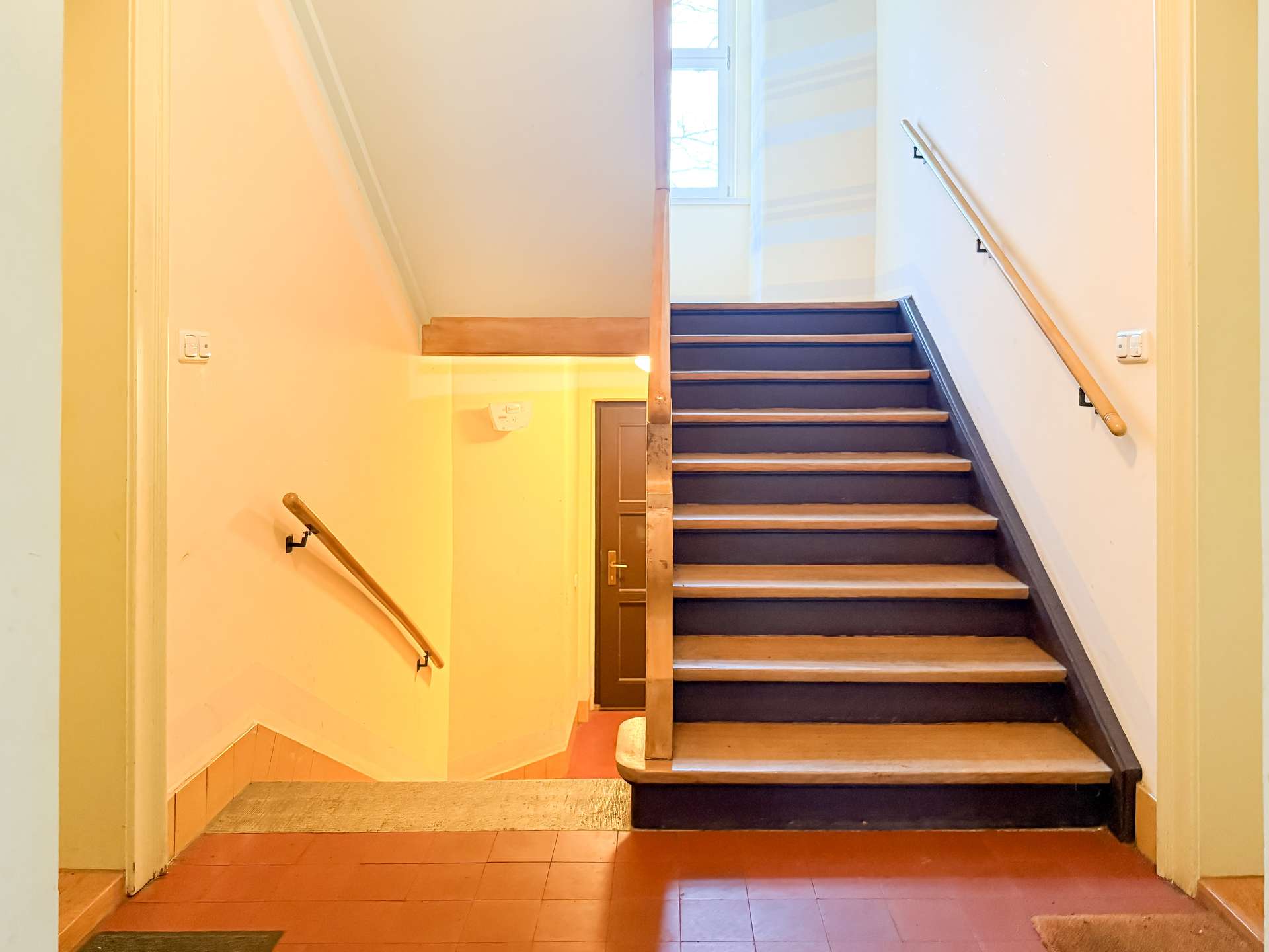Staircase in a building: wooden steps with dark risers, beige walls, and handrails on both sides, leading to a door and a window above.