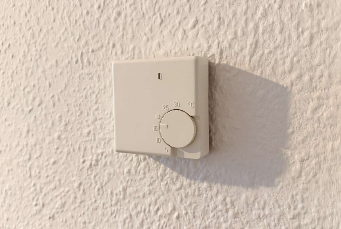 White square thermostat with a circular dial mounted on a textured wall, the dial set to a mid-range temperature.