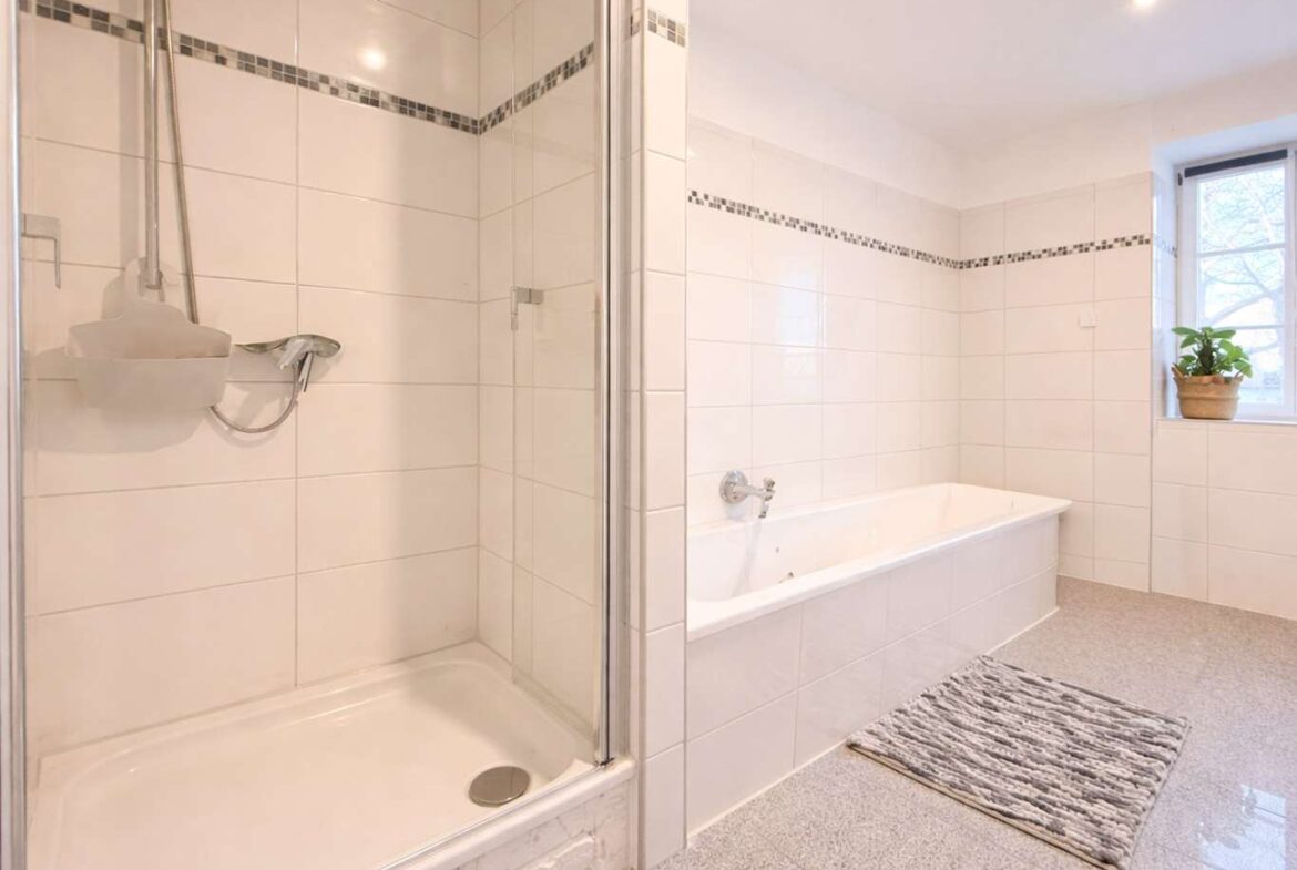 White-tiled bathroom with a glass-enclosed shower on the left and a built-in bathtub on the right, mosaic tile trim, and a window with a potted plant.