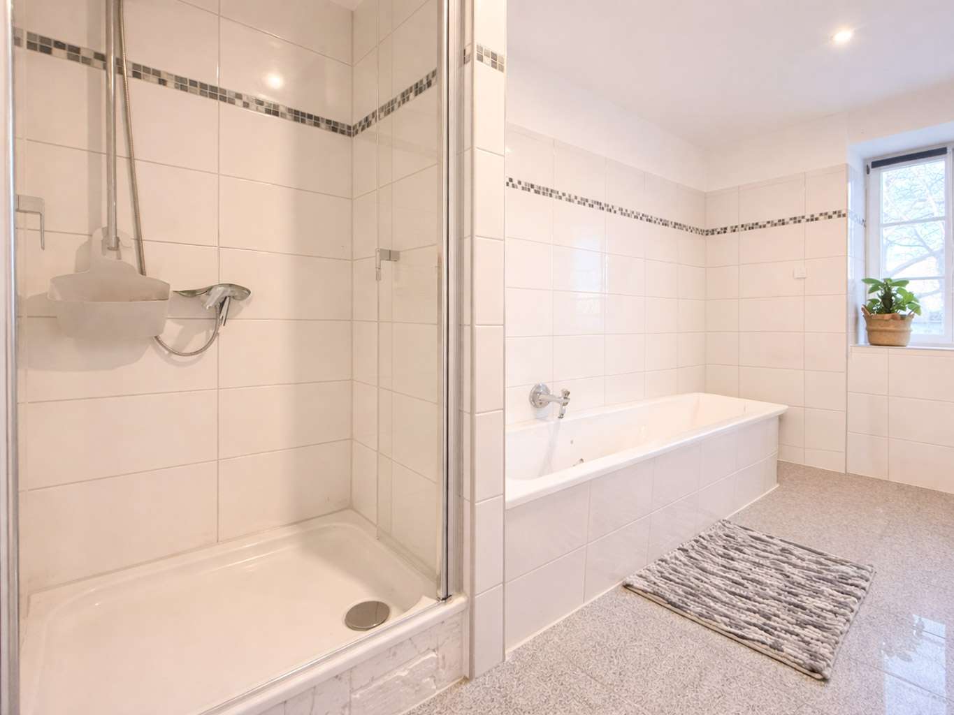 White-tiled bathroom with a glass-enclosed shower on the left and a built-in bathtub on the right, mosaic tile trim, and a window with a potted plant.