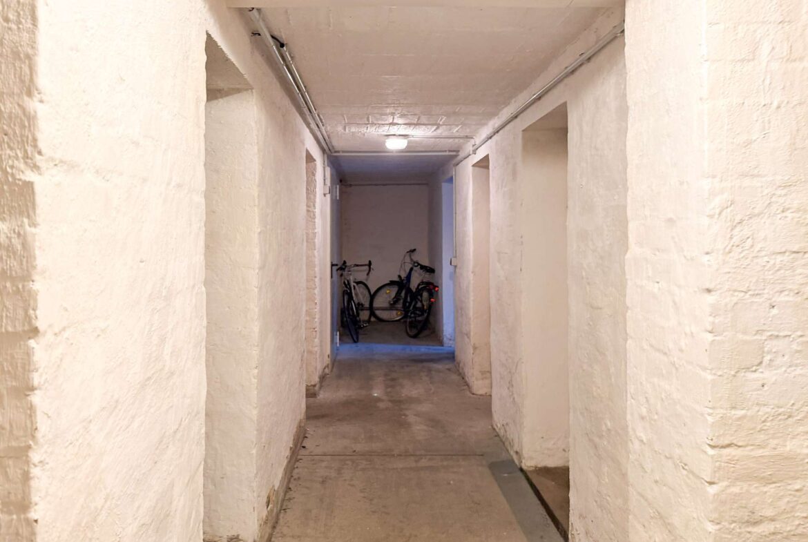 Long concrete basement hallway with whitewashed walls, ending in a small bike storage area. Two bicycles lean against the back wall.