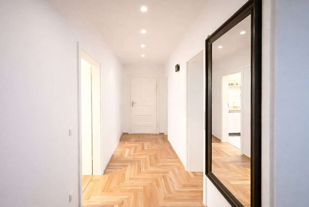 Bright white hallway with a herringbone wood floor, recessed ceiling lights, and a large dark-framed mirror on the right reflecting the corridor ahead.