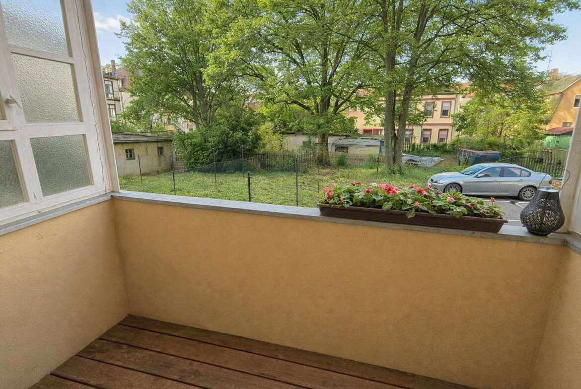 Balcony with a wooden floor and a rectangular planter of pink flowers overlooking a grassy yard and a parked car.