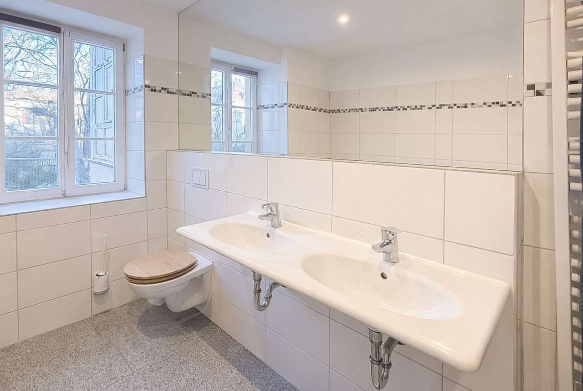 Bright bathroom with a wall-mounted double sink, chrome faucets, a large mirror, and a wall-hung toilet with a wooden seat beside a window.