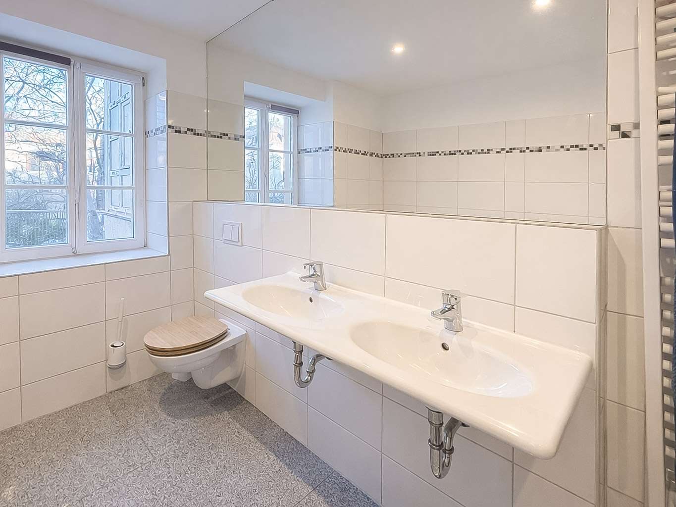 Bright bathroom with a wall-mounted double sink, chrome faucets, a large mirror, and a wall-hung toilet with a wooden seat beside a window.