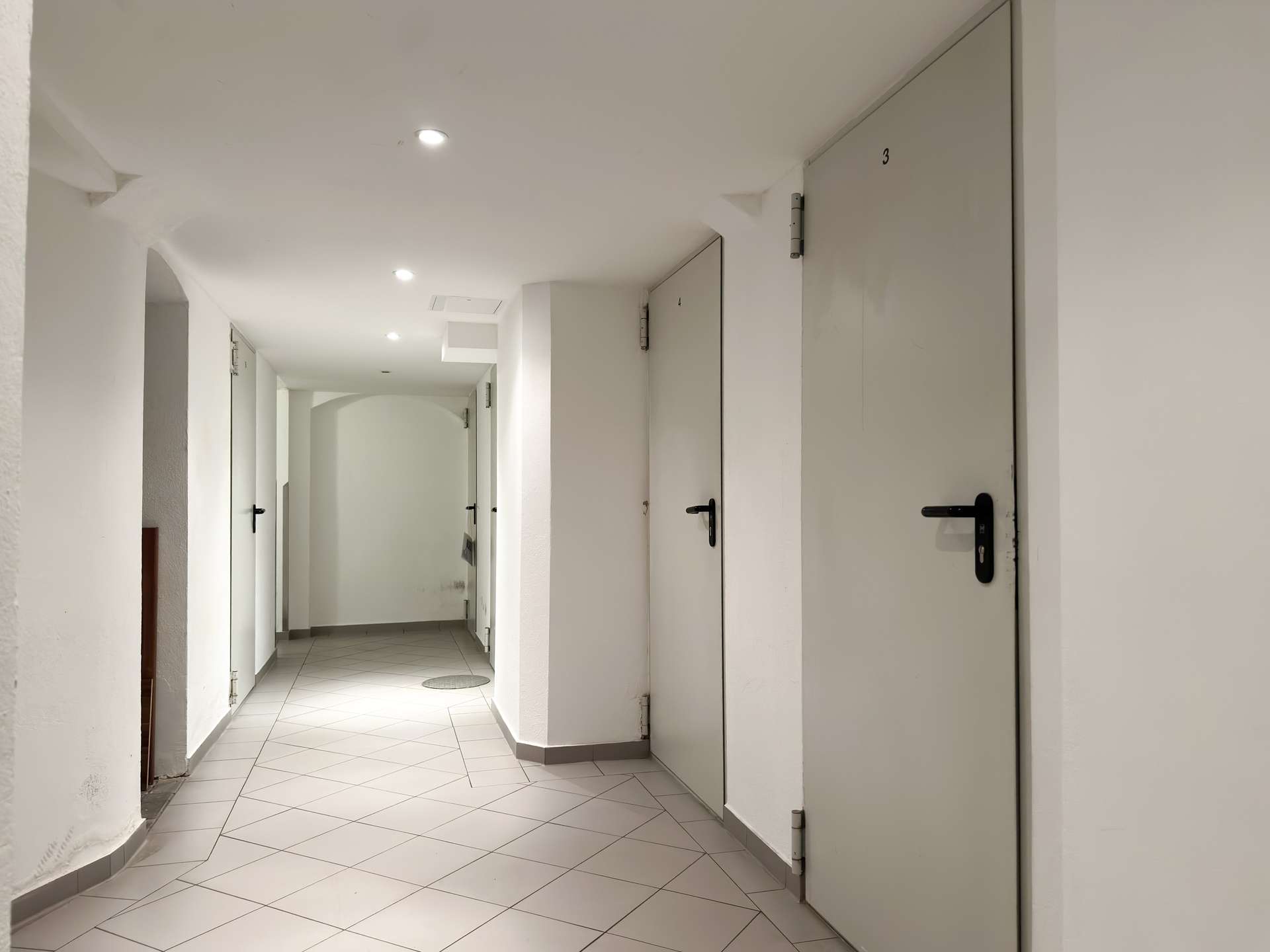 White, well-lit corridor with numbered gray doors along the right side and a tiled floor in a residential-looking building.