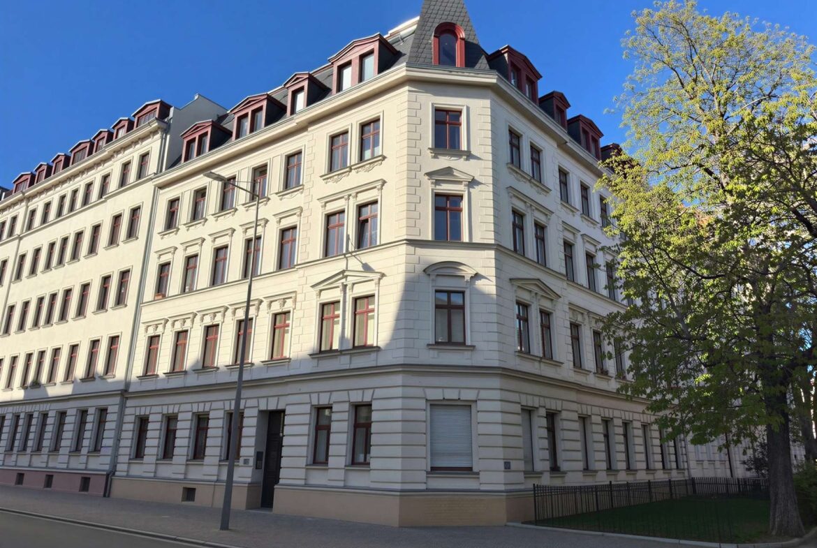 Corner view of a white ornate apartment building with red window frames and a steep mansard roof against a blue sky.