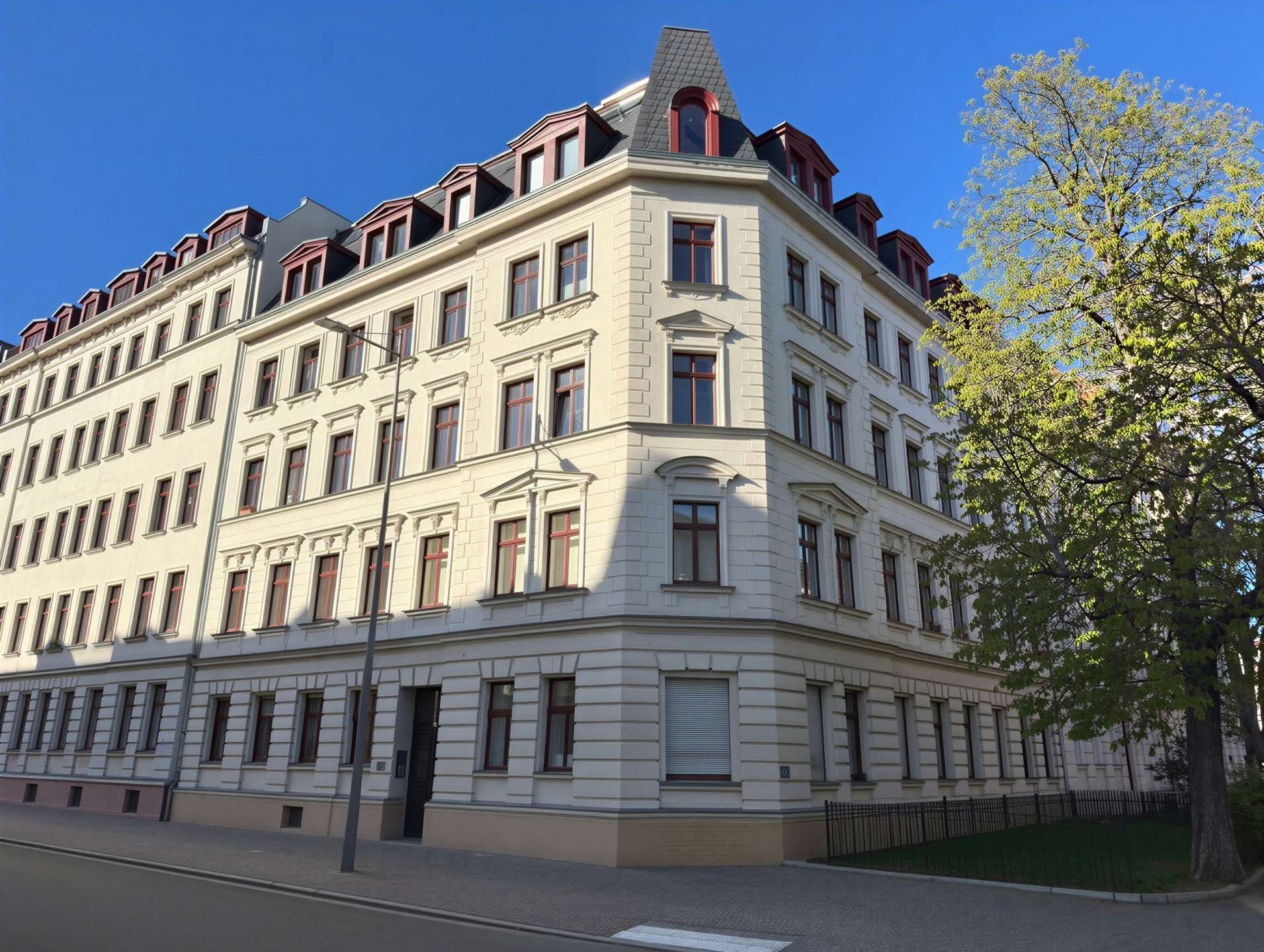 Corner view of a white ornate apartment building with red window frames and a steep mansard roof against a blue sky.