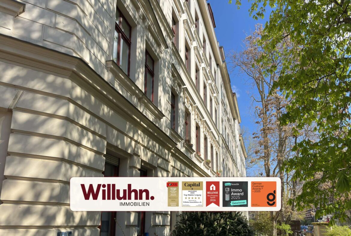 Cream-colored, multi-story building with decorative cornices and red-framed windows; foreground shows a Willuhn Immobilien sign with award badges under a blue sky and trees.