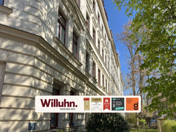 Cream-colored, multi-story building with decorative cornices and red-framed windows; foreground shows a Willuhn Immobilien sign with award badges under a blue sky and trees.