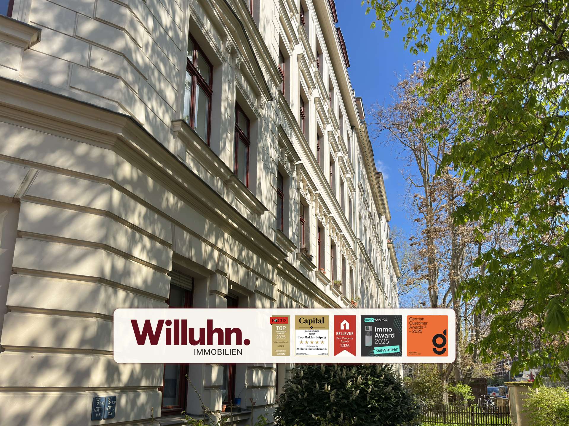 Cream-colored, multi-story building with decorative cornices and red-framed windows; foreground shows a Willuhn Immobilien sign with award badges under a blue sky and trees.
