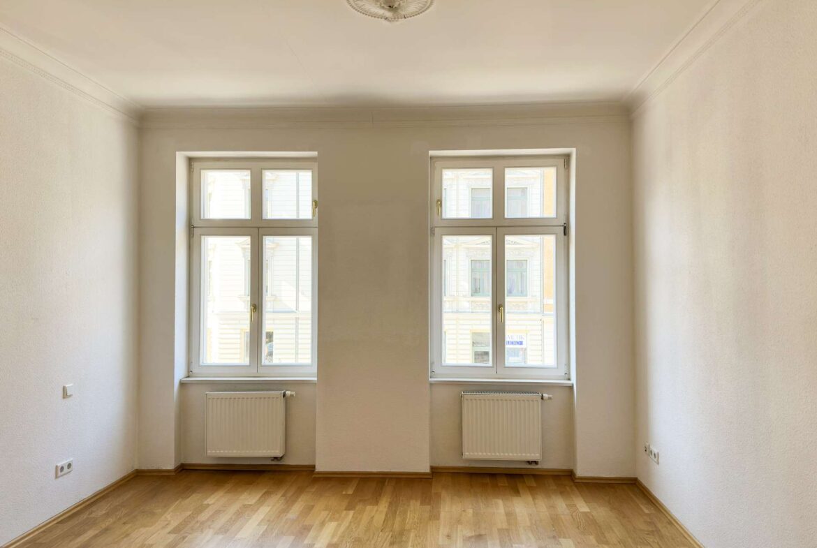 Empty rectangular room with two tall windows, white walls, and light hardwood flooring; radiators beneath the windows.