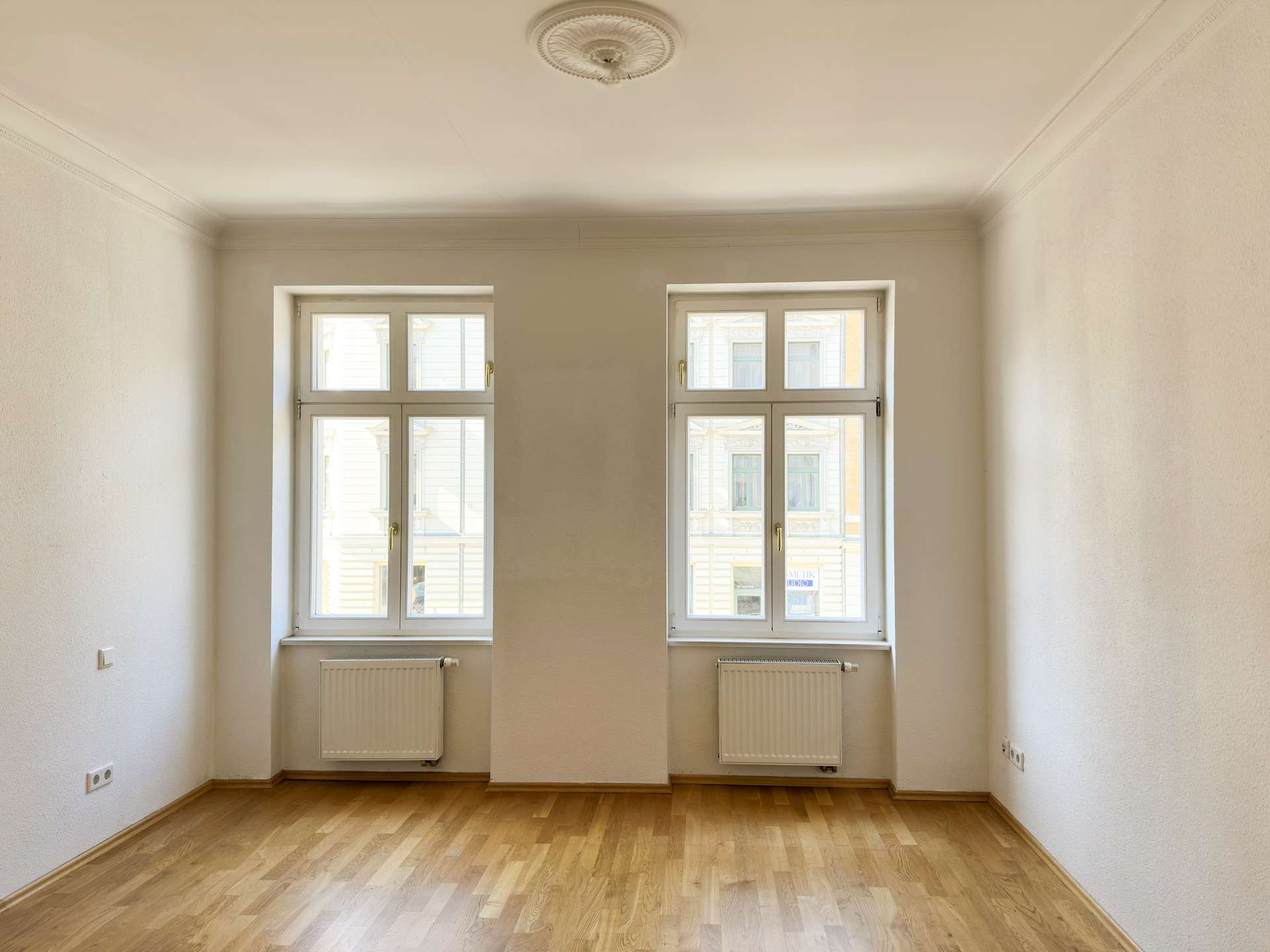 Empty rectangular room with two tall windows, white walls, and light hardwood flooring; radiators beneath the windows.
