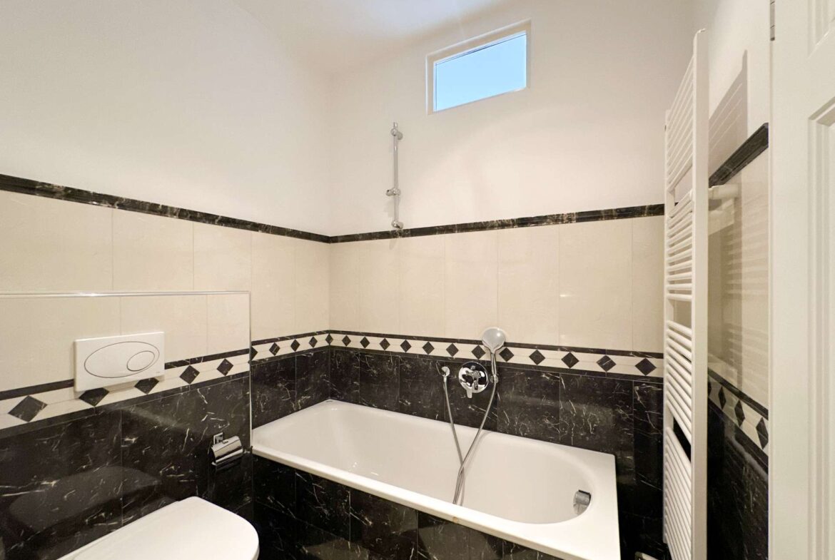 White bathroom with a rectangular bathtub, handheld shower, and black marble tiling along the lower walls; a slim vertical towel warmer on the right and a small high window above.