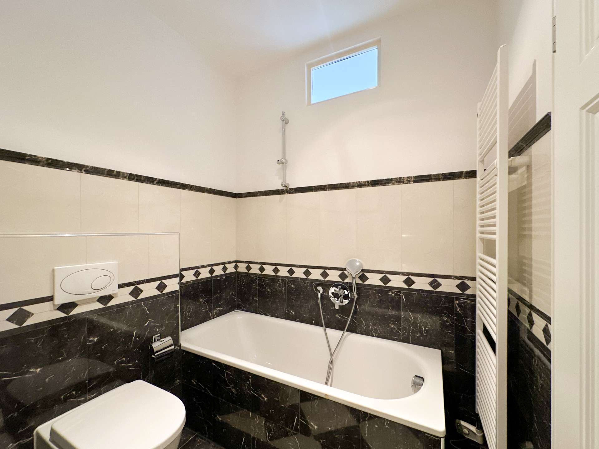 White bathroom with a rectangular bathtub, handheld shower, and black marble tiling along the lower walls; a slim vertical towel warmer on the right and a small high window above.