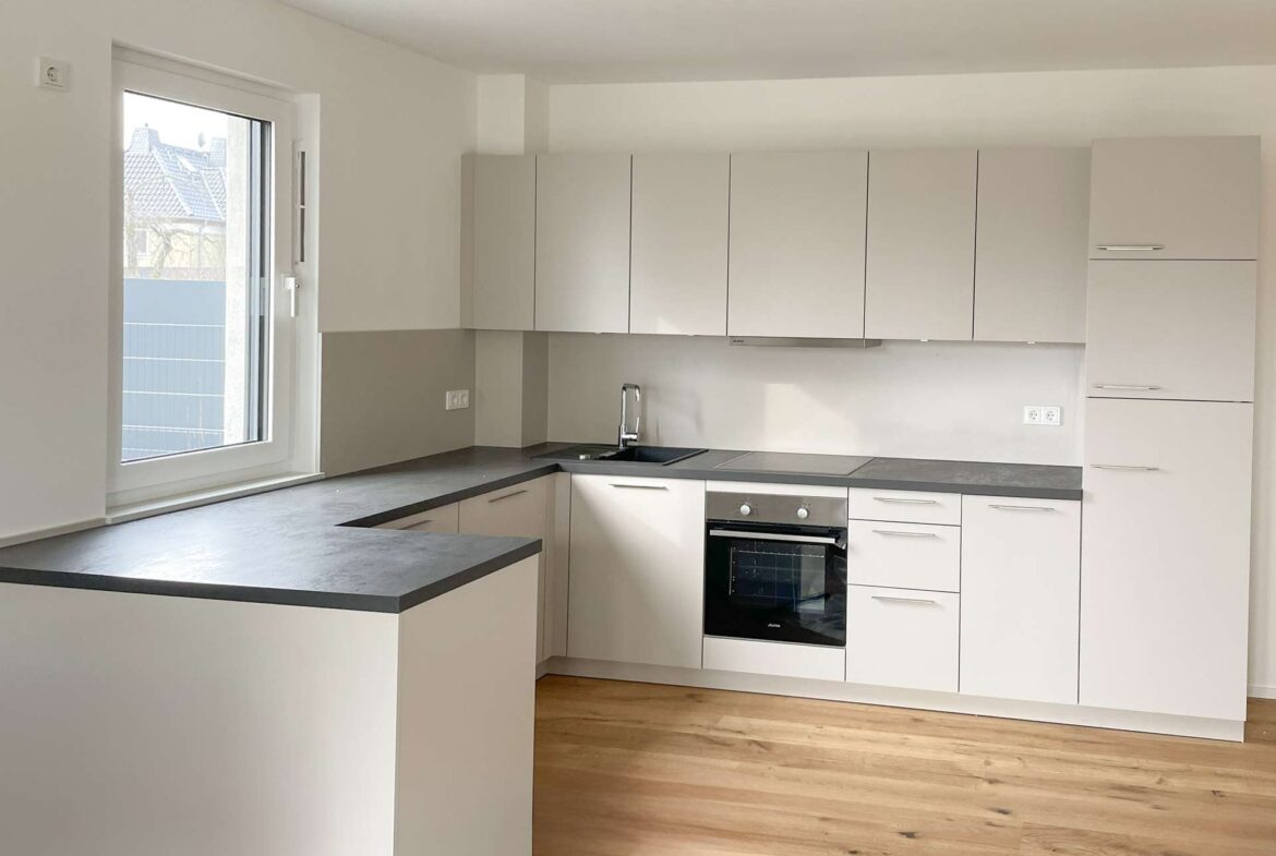 Modern L-shaped kitchen with white cabinets, grey countertops, and a built-in oven under a windowed wall. The light wood floor runs across the room.