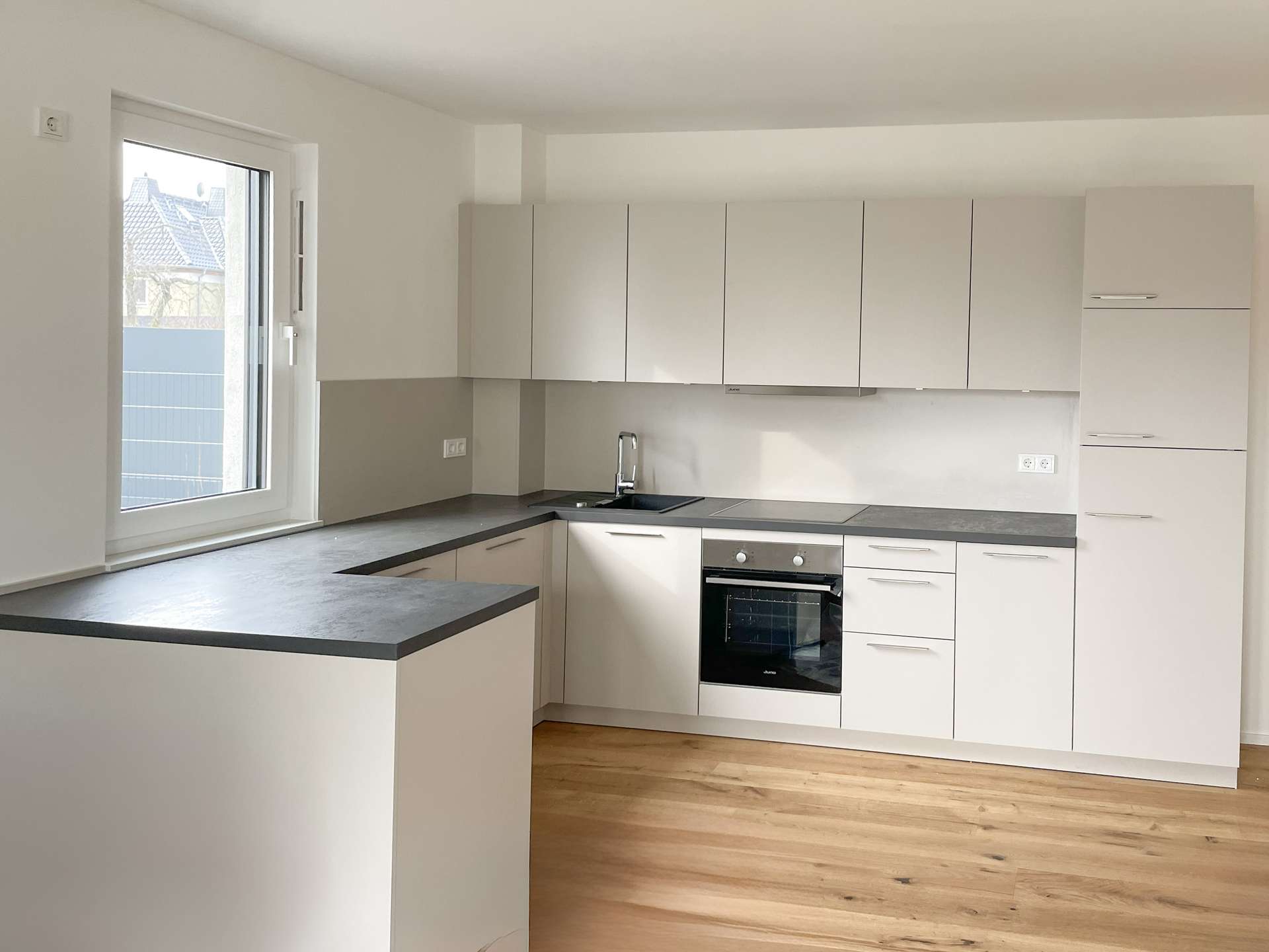 Modern L-shaped kitchen with white cabinets, grey countertops, and a built-in oven under a windowed wall. The light wood floor runs across the room.