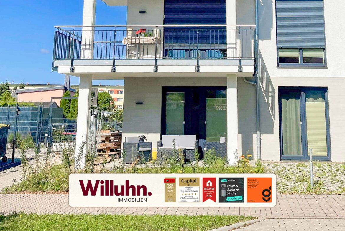 Front of a modern two-story building with a patio and large glass doors; a Willuhn Immobilien signboard with award logos is in the foreground.