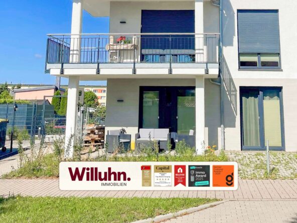 Front of a modern two-story building with a patio and large glass doors; a Willuhn Immobilien signboard with award logos is in the foreground.