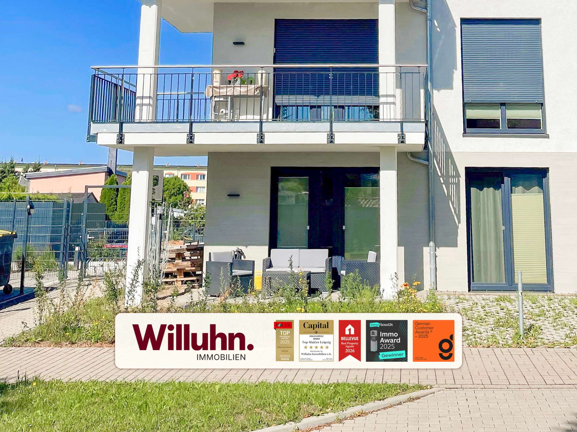 Front of a modern two-story building with a patio and large glass doors; a Willuhn Immobilien signboard with award logos is in the foreground.