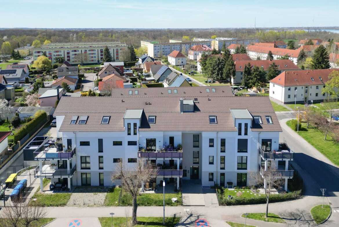 Aerial view of a modern white apartment building with red-tiled roofs, neighboring houses, trees, and a street in a suburban neighborhood under a blue sky.
