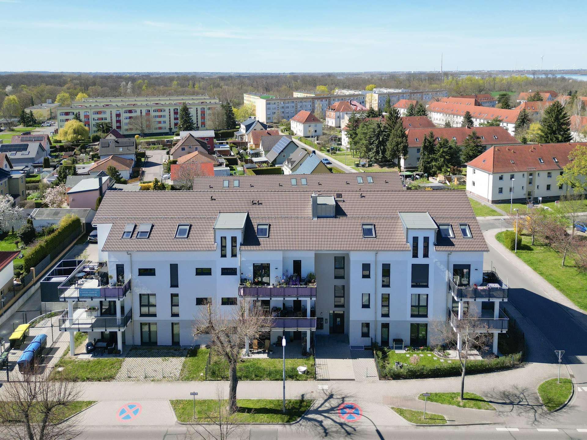 Aerial view of a modern white apartment building with red-tiled roofs, neighboring houses, trees, and a street in a suburban neighborhood under a blue sky.
