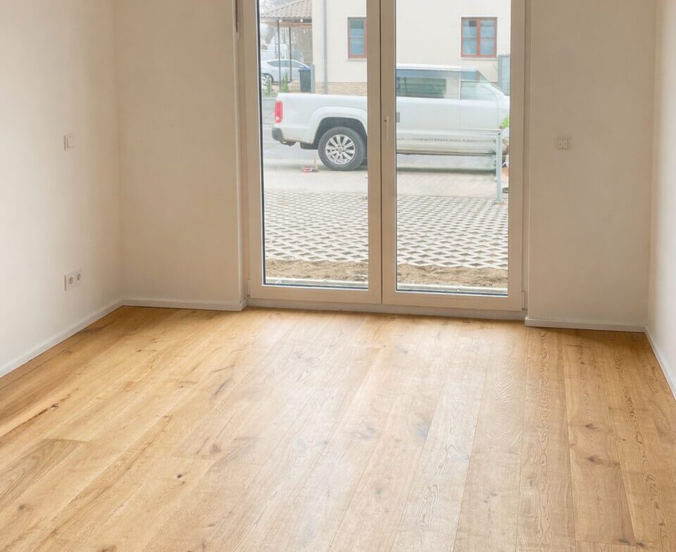 Empty room with light wood floor and large glass door leading to an outside area, white walls and minimal fixtures inside.