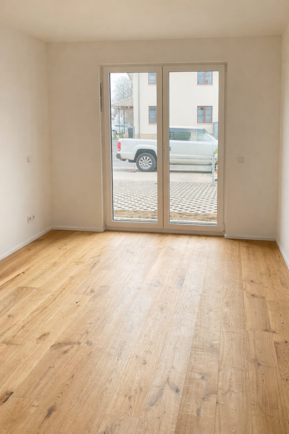 Empty room with light wood floor and large glass door leading to an outside area, white walls and minimal fixtures inside.