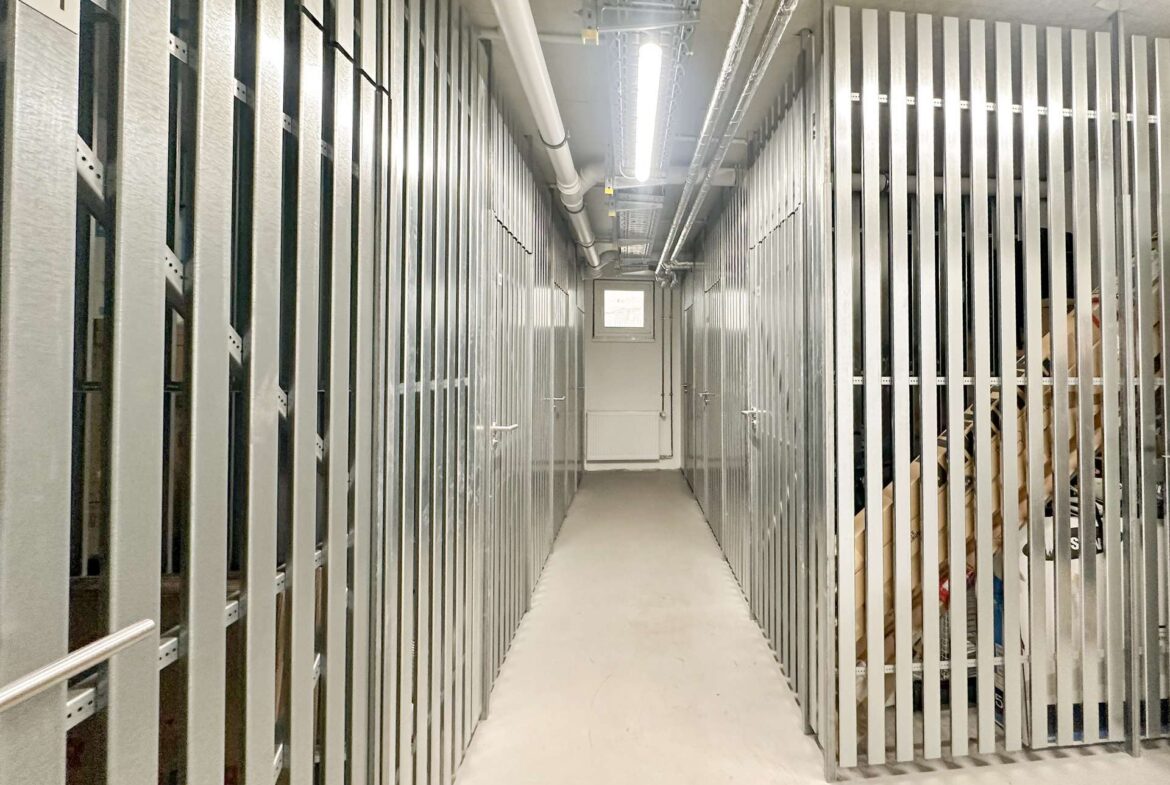 Narrow corridor in a storage facility with tall metal rack rails on both sides and overhead pipes, illuminated by a fluorescent light.
