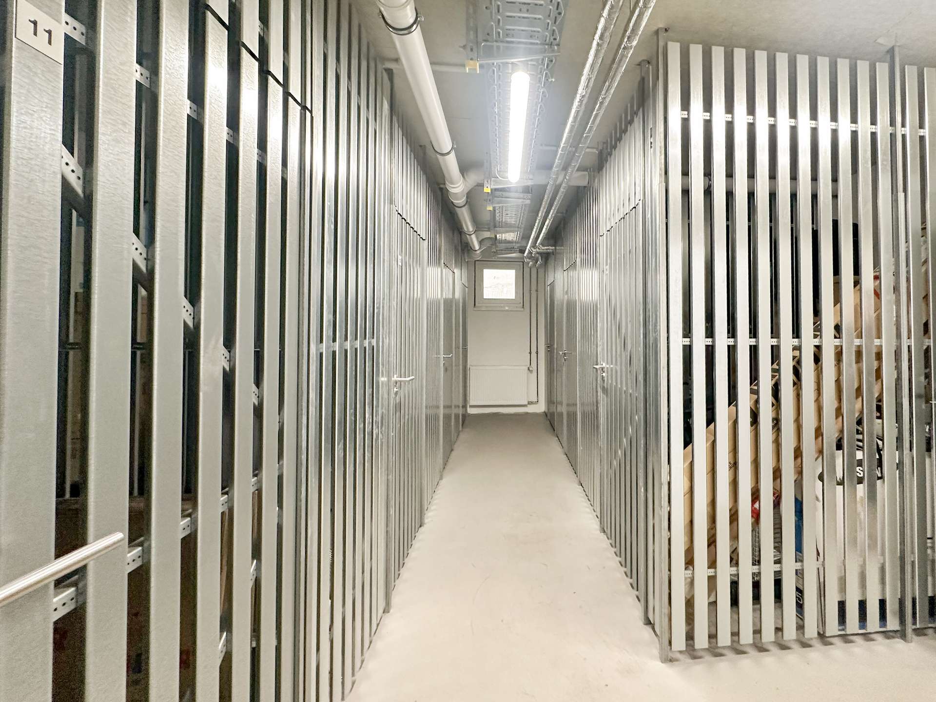 Narrow corridor in a storage facility with tall metal rack rails on both sides and overhead pipes, illuminated by a fluorescent light.