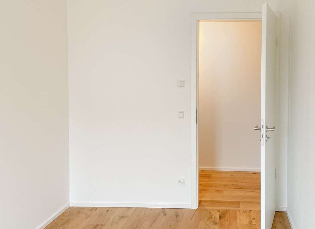 Empty white room with a light wooden floor and an open white door revealing a brighter adjacent space