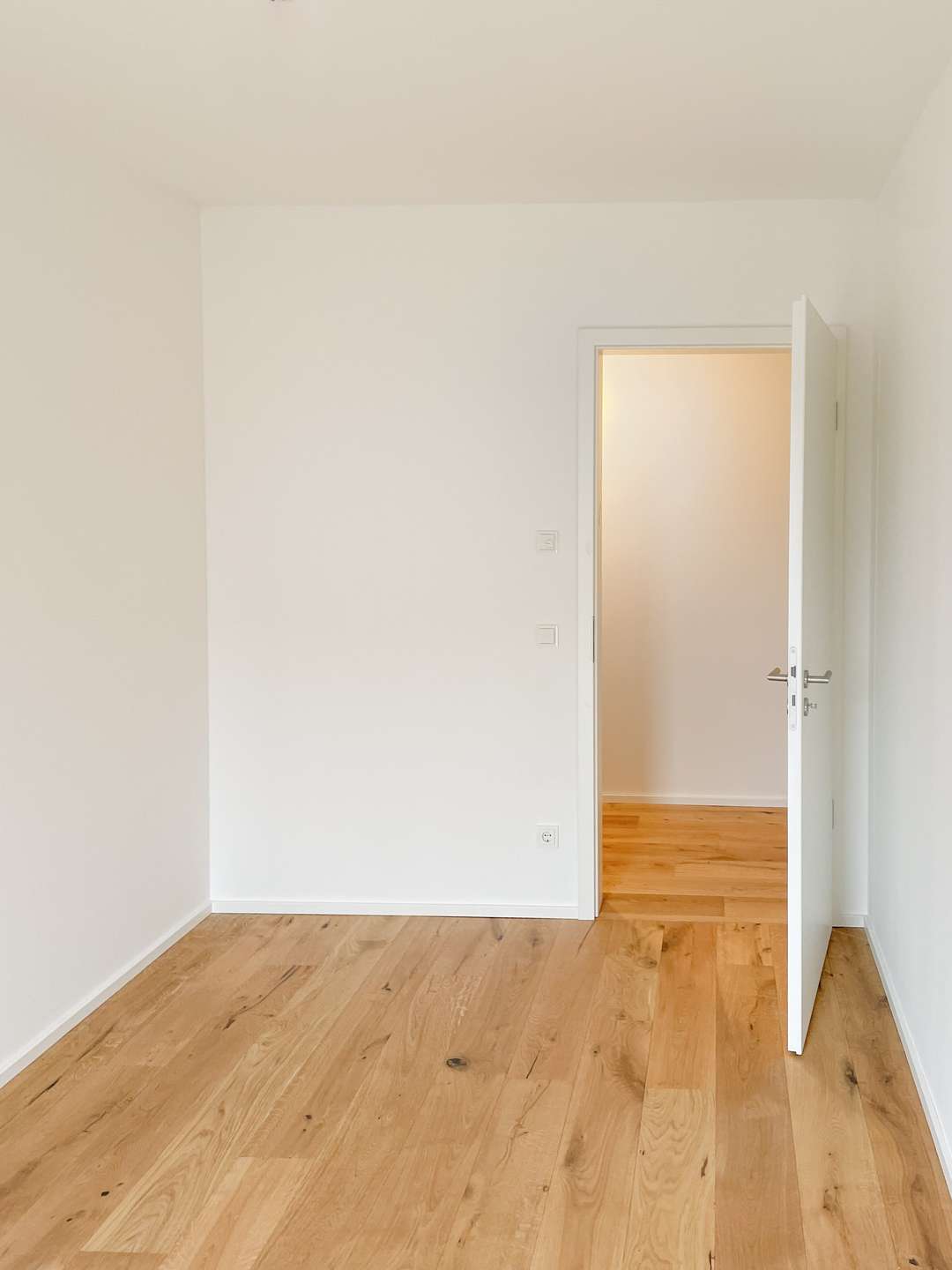 Empty white room with a light wooden floor and an open white door revealing a brighter adjacent space
