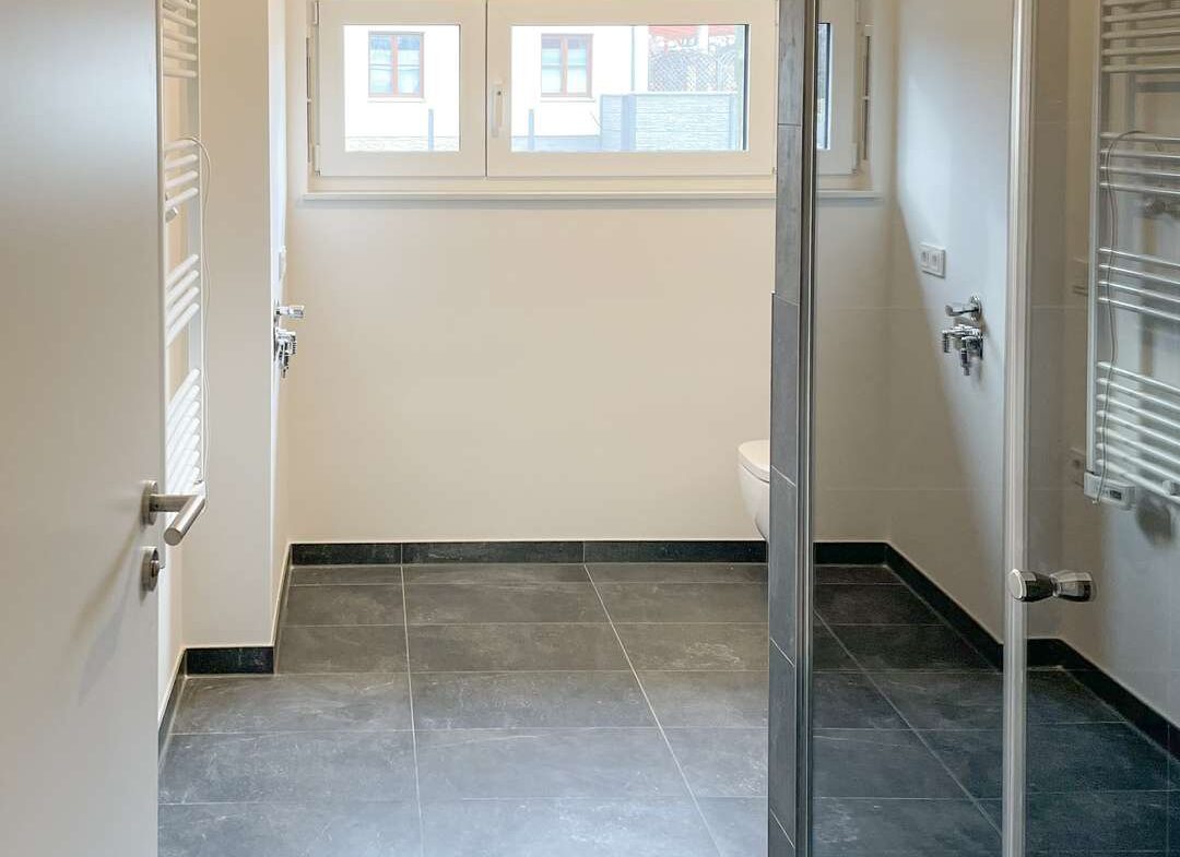 Bright minimalist bathroom with gray floor tiles, a glass shower enclosure on the right, a towel radiator on the left, and a horizontal window.