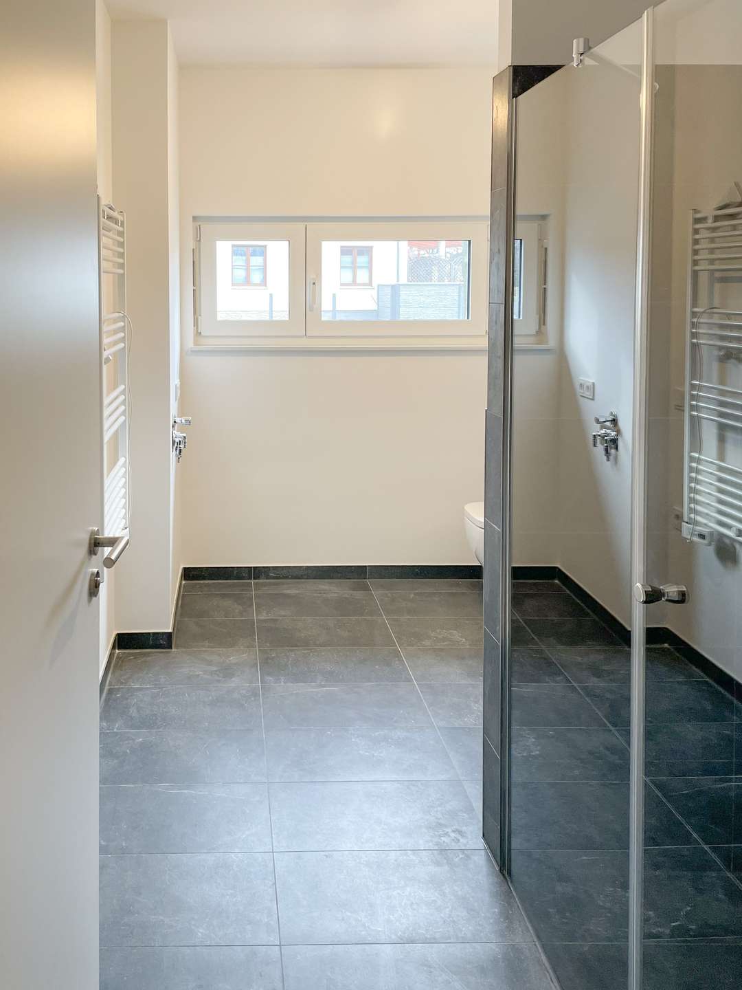 Bright minimalist bathroom with gray floor tiles, a glass shower enclosure on the right, a towel radiator on the left, and a horizontal window.