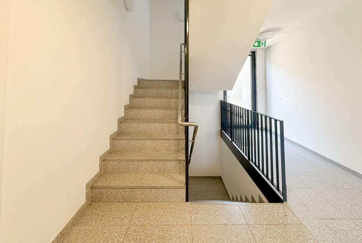 Stairwell inside a building with beige speckled stairs, metal handrail, and a railing along a corridor; exit sign visible near the door upstairs.