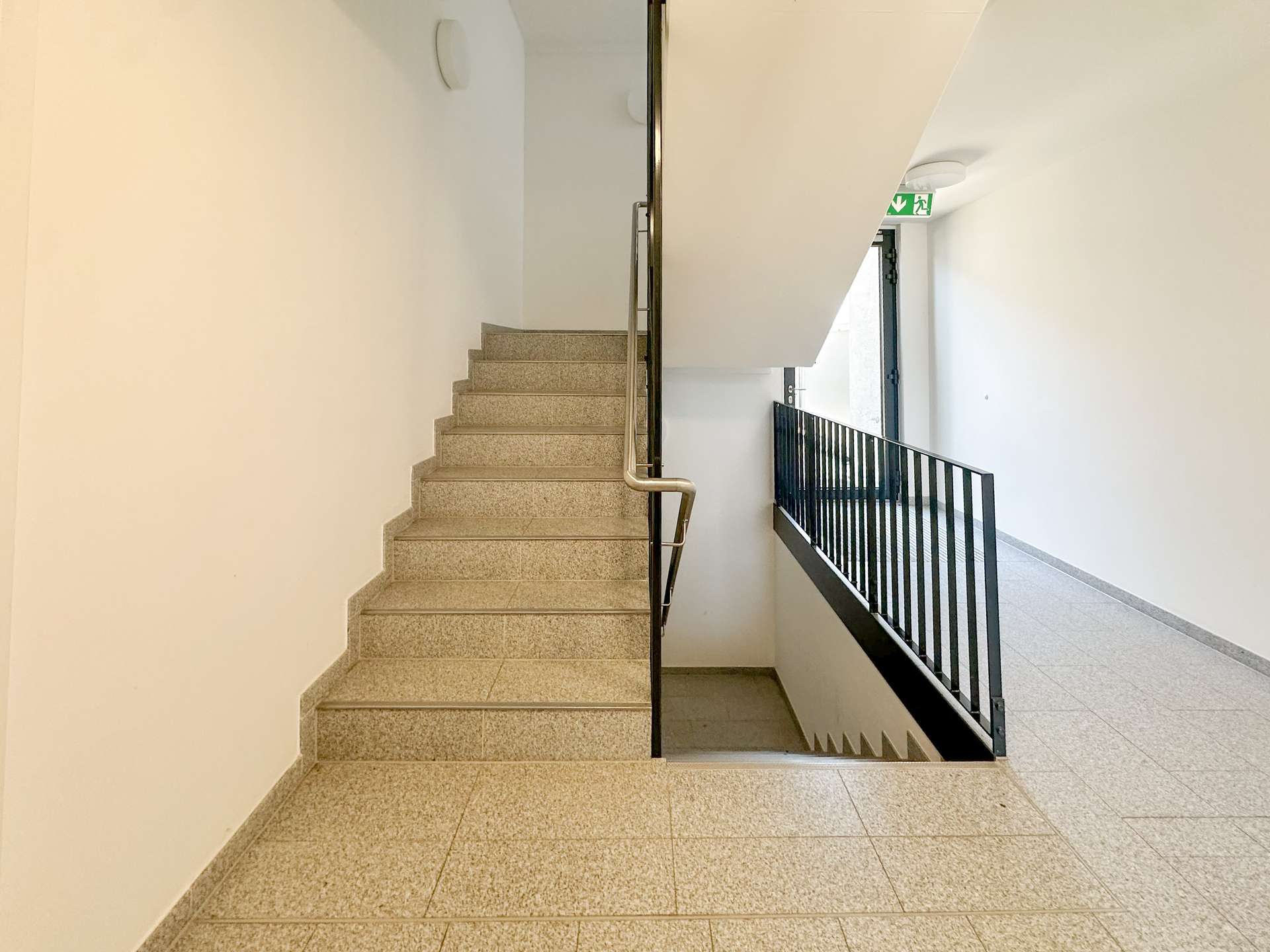 Stairwell inside a building with beige speckled stairs, metal handrail, and a railing along a corridor; exit sign visible near the door upstairs.