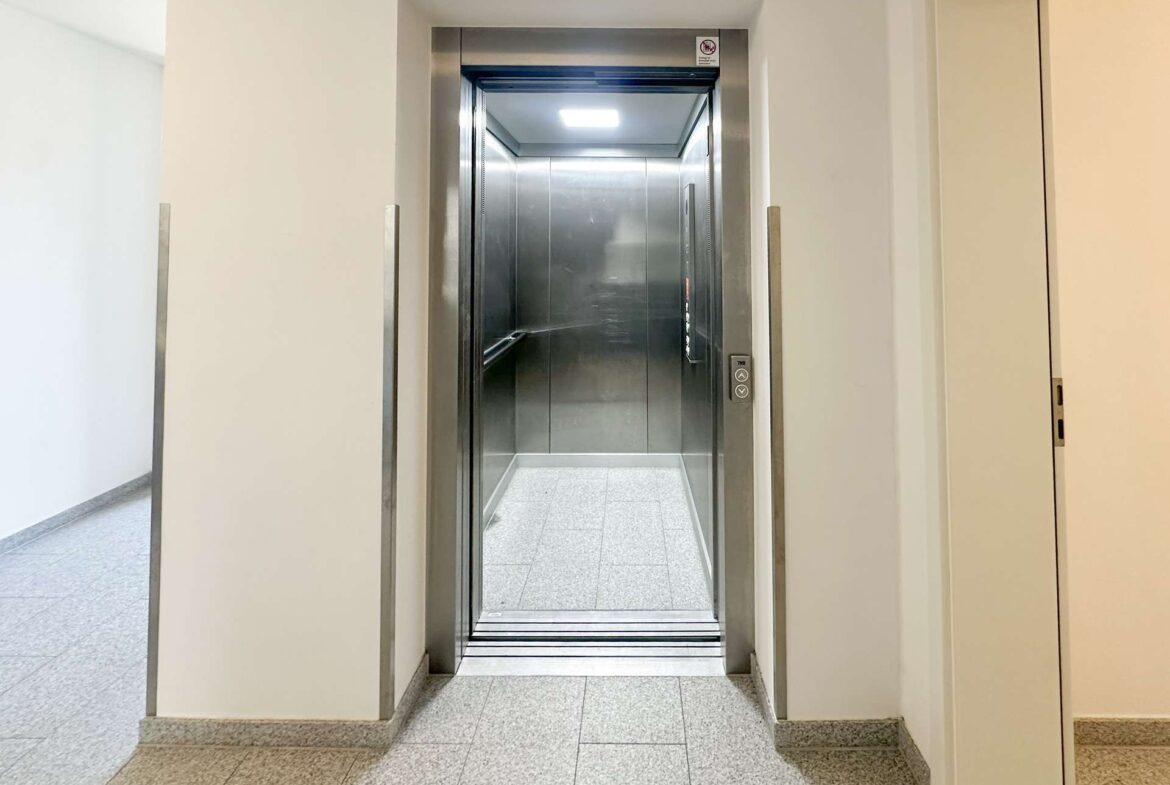 Elevator with stainless steel doors open in a light hallway, showing a clean, modern lobby area behind it.