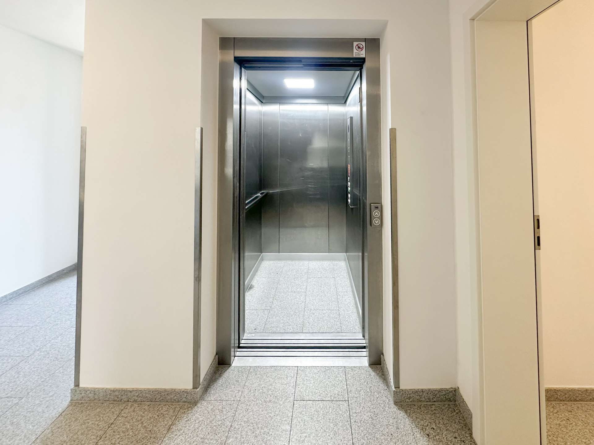 Elevator with stainless steel doors open in a light hallway, showing a clean, modern lobby area behind it.