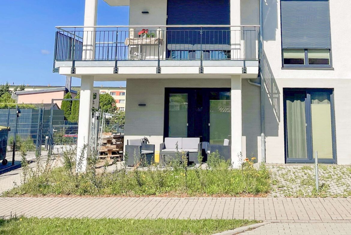Modern two-story building with a balcony and outdoor seating, blue shuttered windows, and a paved path in the foreground.