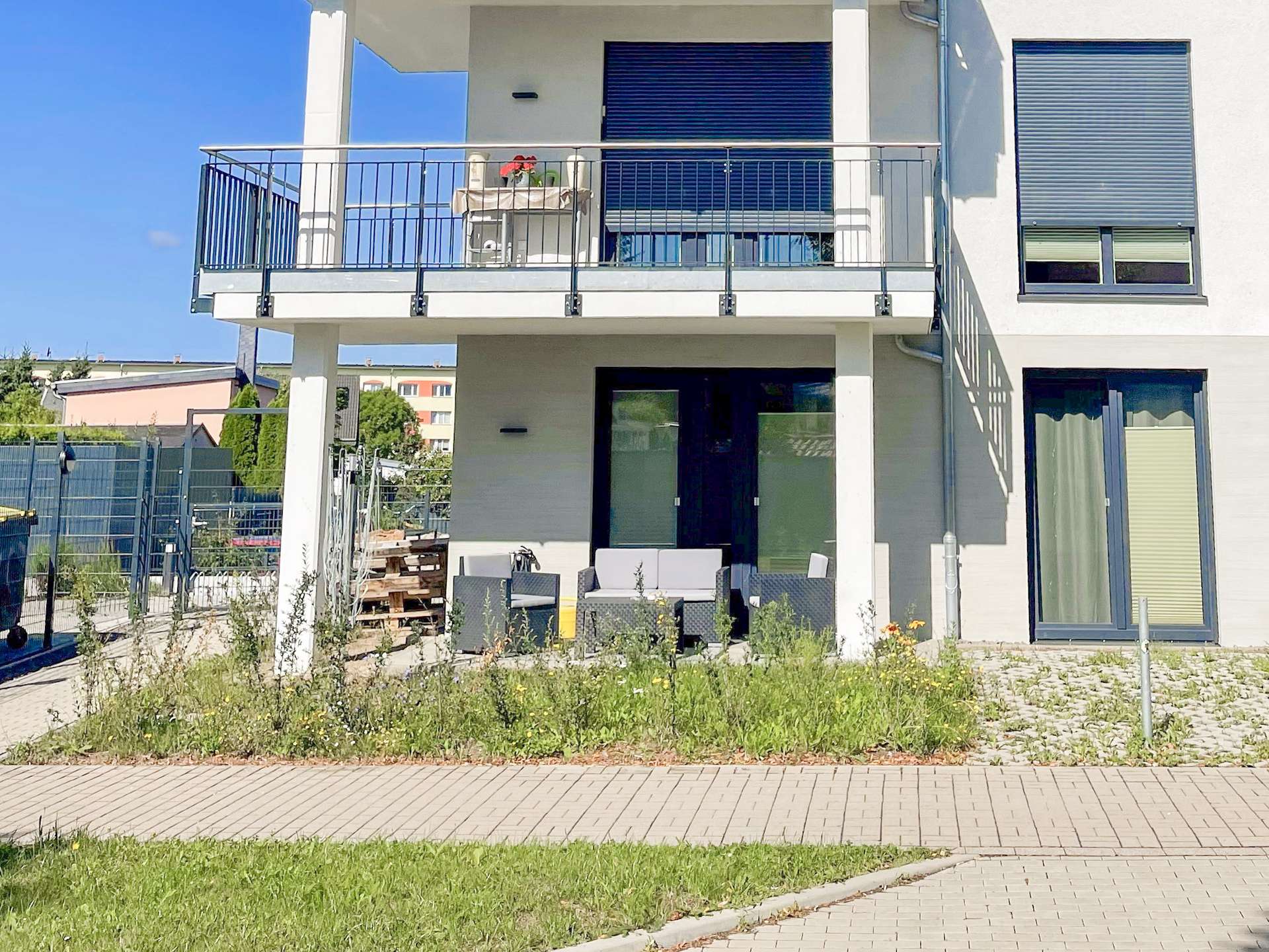 Modern two-story building with a balcony and outdoor seating, blue shuttered windows, and a paved path in the foreground.