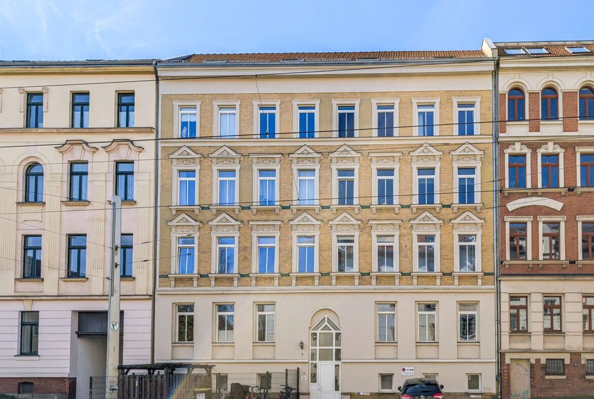 Row of ornate beige and brick townhouses with tall sash windows along a street; a car and bicycles parked in front.