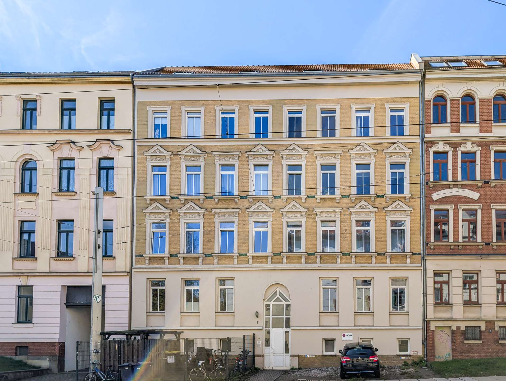 Row of ornate beige and brick townhouses with tall sash windows along a street; a car and bicycles parked in front.